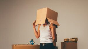 Persoon met een verhuisdoos over het hoofd tijdens zelf verhuizen, omringd door dozen en verhuisspullen